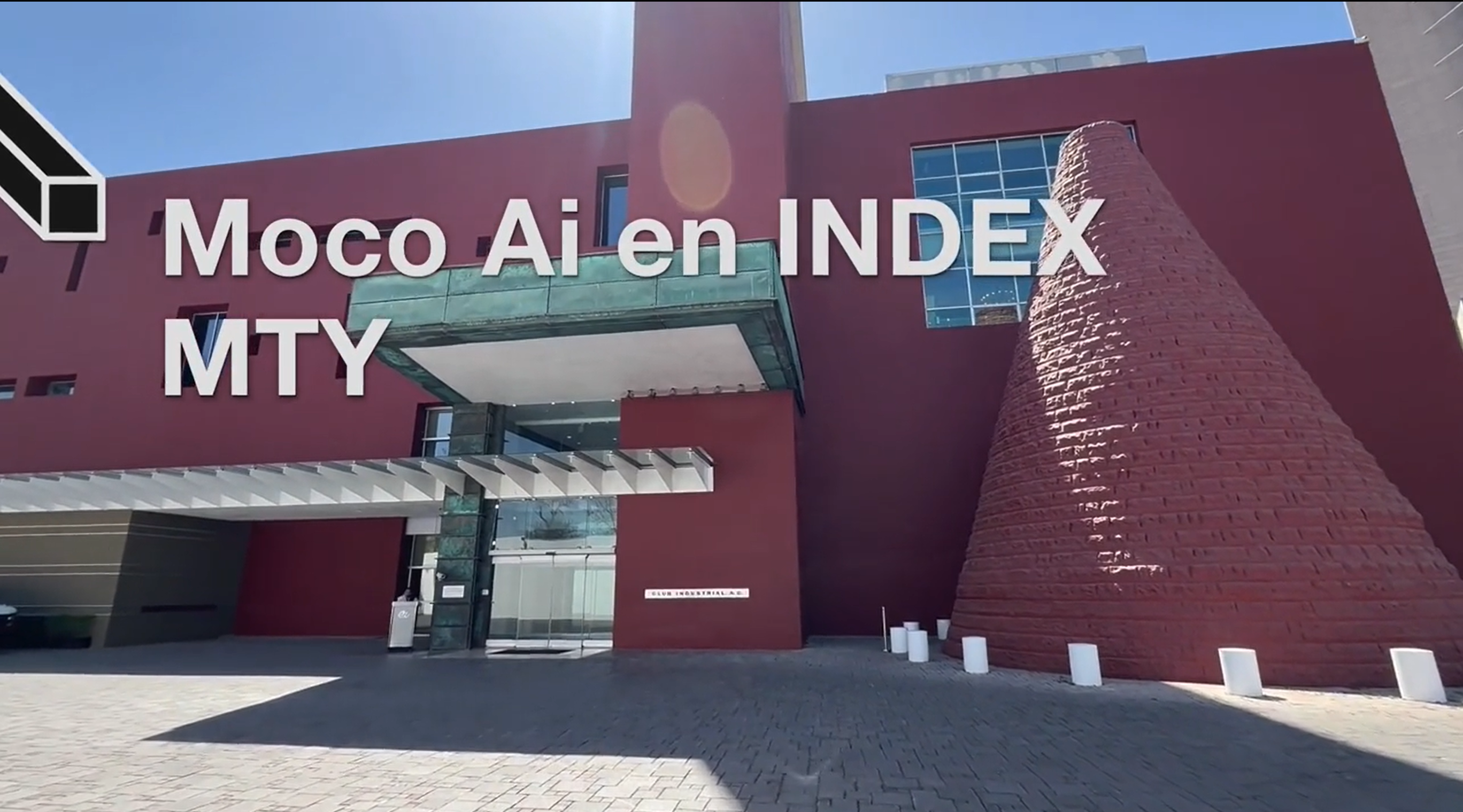Fachada de un edificio moderno de color rojo con detalles en cristal y una estructura cónica de ladrillo. En la imagen aparece el texto 'Moco AI en INDEX MTY
